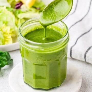 Close up of a spoon drizzling fresh homemade basil vinaigrette in a glass jar with a lemon and salad in the background.