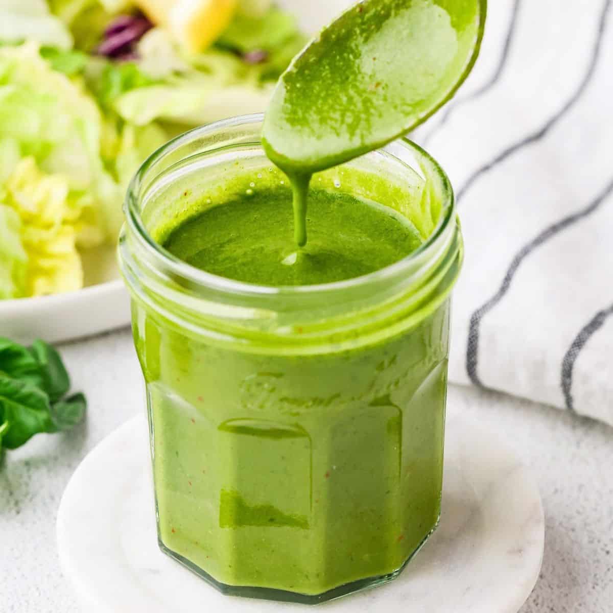 Close up of a spoon drizzling fresh homemade basil vinaigrette in a glass jar with a lemon and salad in the background.
