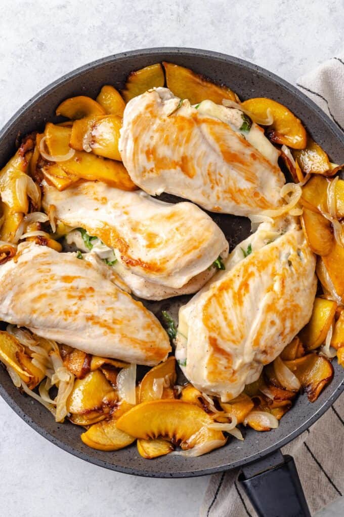 Seared brie cheese stuffed chicken breasts in a skillet with sautéed slices of onion and peaches.