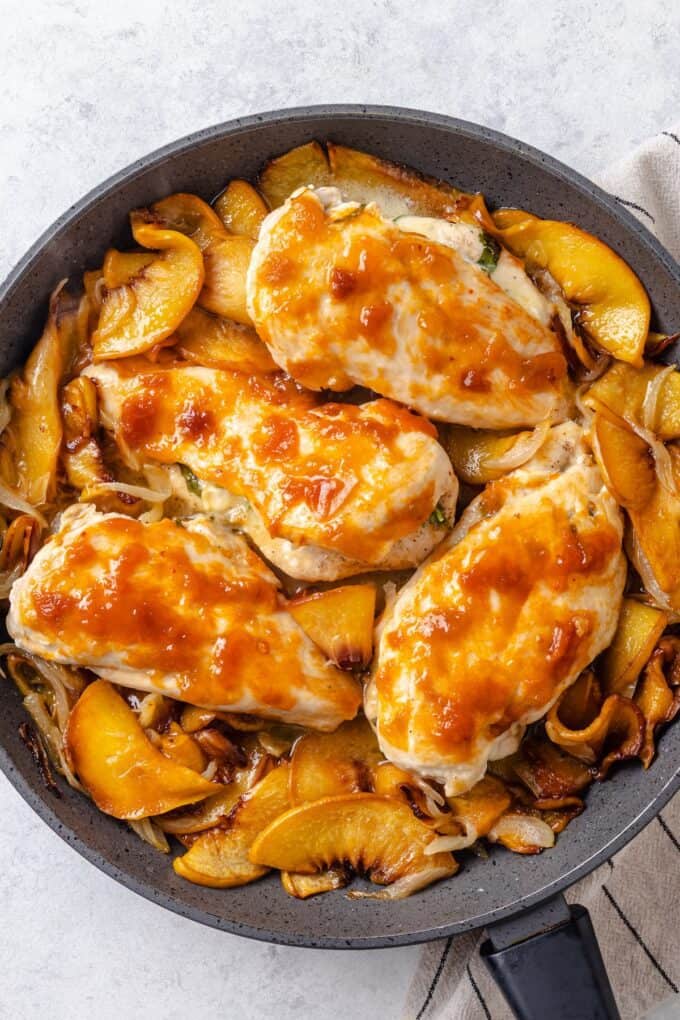 Seared brie cheese stuffed chicken breasts brushed with peach preserves in a skillet with sautéed slices of onion and peaches.