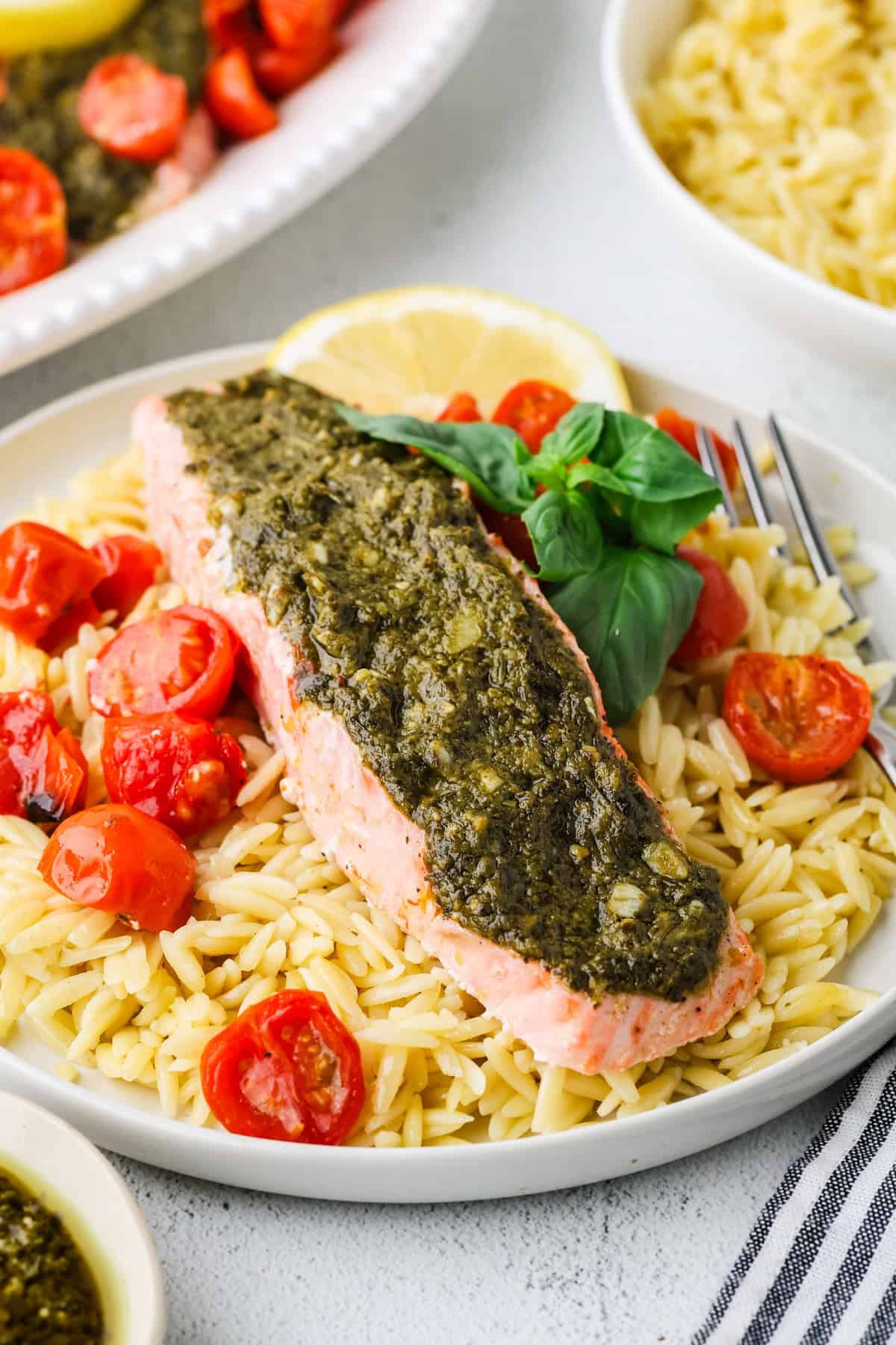 A white dinner plate with a pesto salmon filet atop orzo with blistered tomatoes and basil to the side.