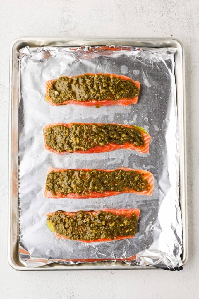 Pesto topped salmon filets on a foil lined baking sheet before being cooked.