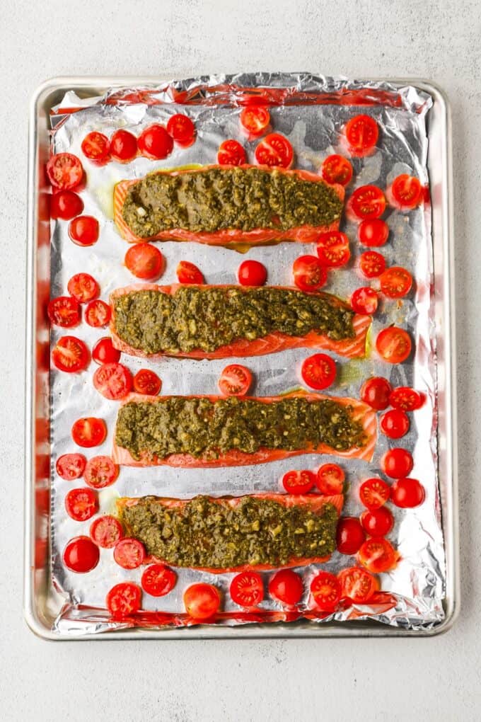 Pesto topped salmon filets and halved cherry tomatoes on a foil lined baking sheet before being cooked.