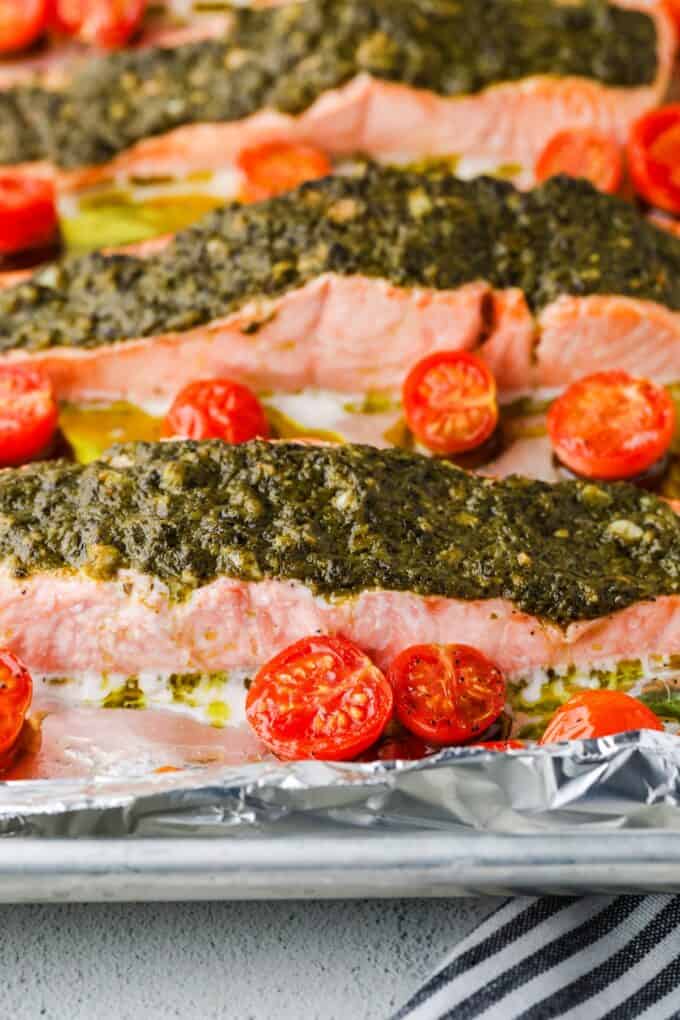 Close up of broiled pesto salmon on a foil lined baking sheet with cherry tomatoes scattered about.