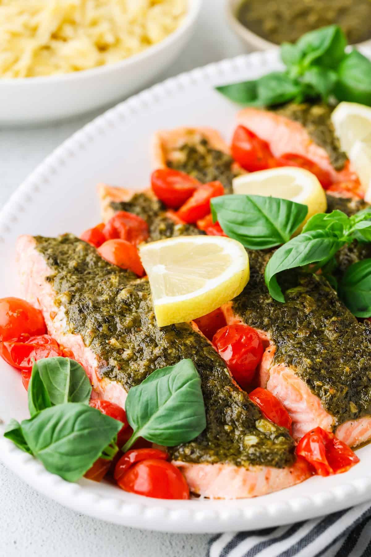 Pesto salmon filets served in an oval serving dish with blistered cherry tomatoes, lemon slices and fresh basil.