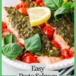 Pesto salmon filets served in an oval serving dish with blistered cherry tomatoes, lemon slices and fresh basil with a green border and text around the image.