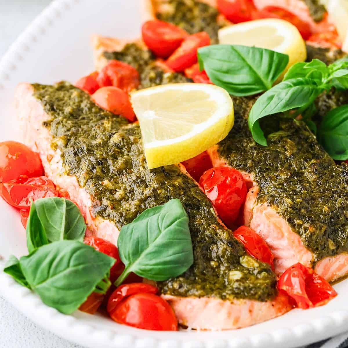 Broiled pesto salmon filets served in an oval serving dish with blistered cherry tomatoes, lemon slices and fresh basil.