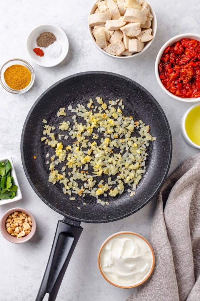 Sautéed onions and garlic in a skillet with bowls of other ingredients around it to be added in.