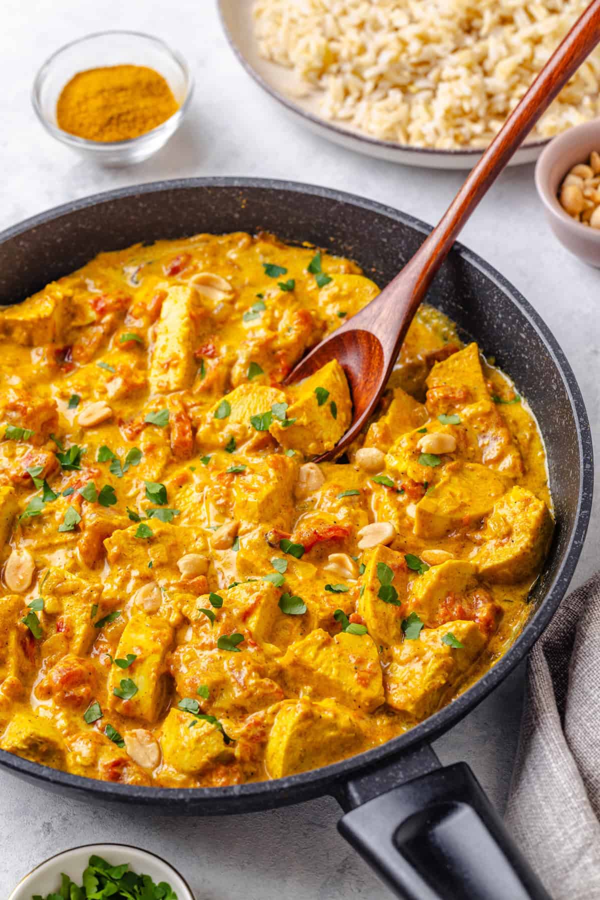 A wooden spoon resting in a skillet filled with a creamy tomato chicken curry topped with peanuts and cilantro.