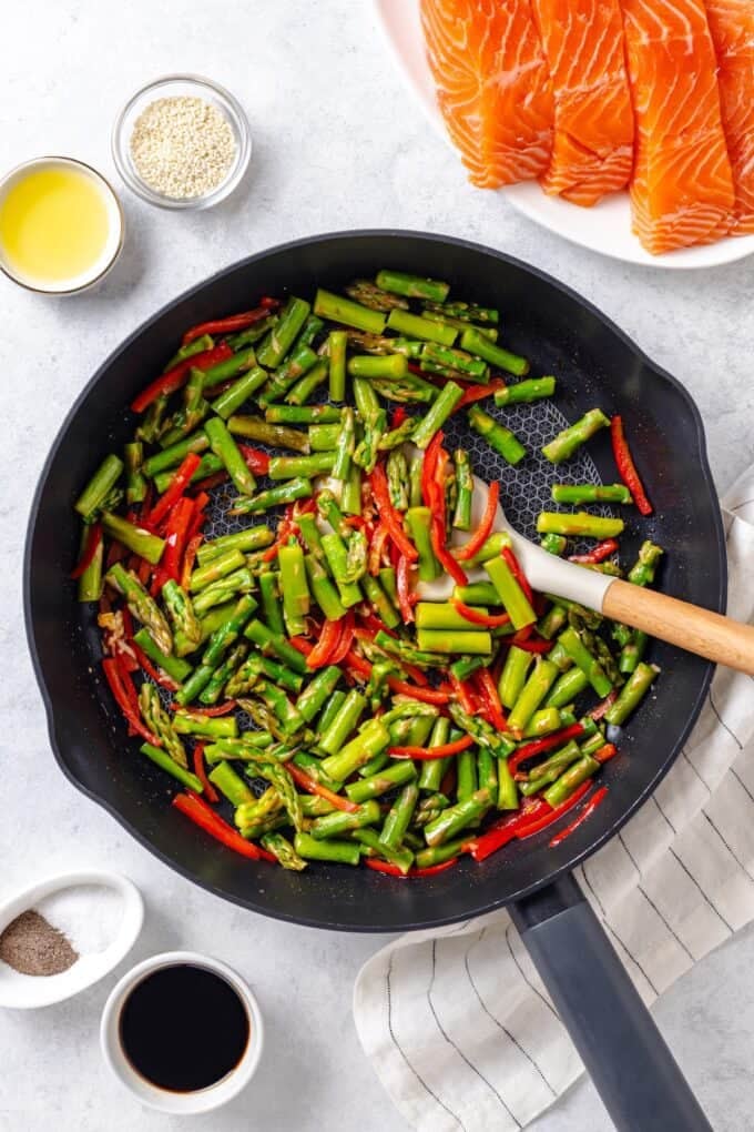 A large skillet with a mixture of cut asparagus and red bell pepper with a spoon resting in the pan.