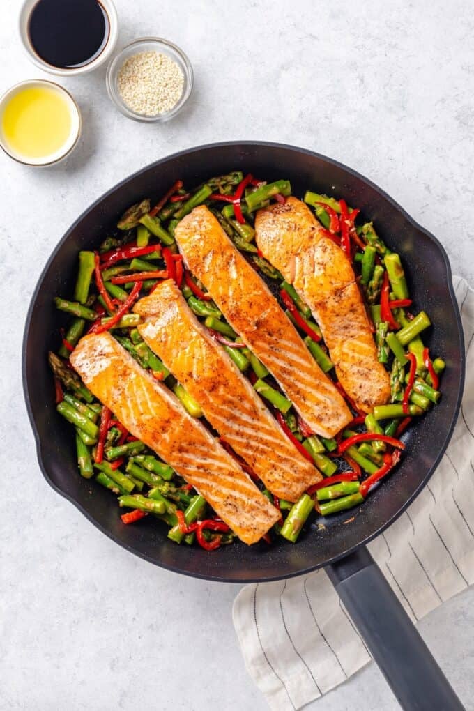 A large dark skillet with seared salmon filets resting on top of a mixture of asparagus and red bell pepper with some sauce ingredients in bowls to the side.