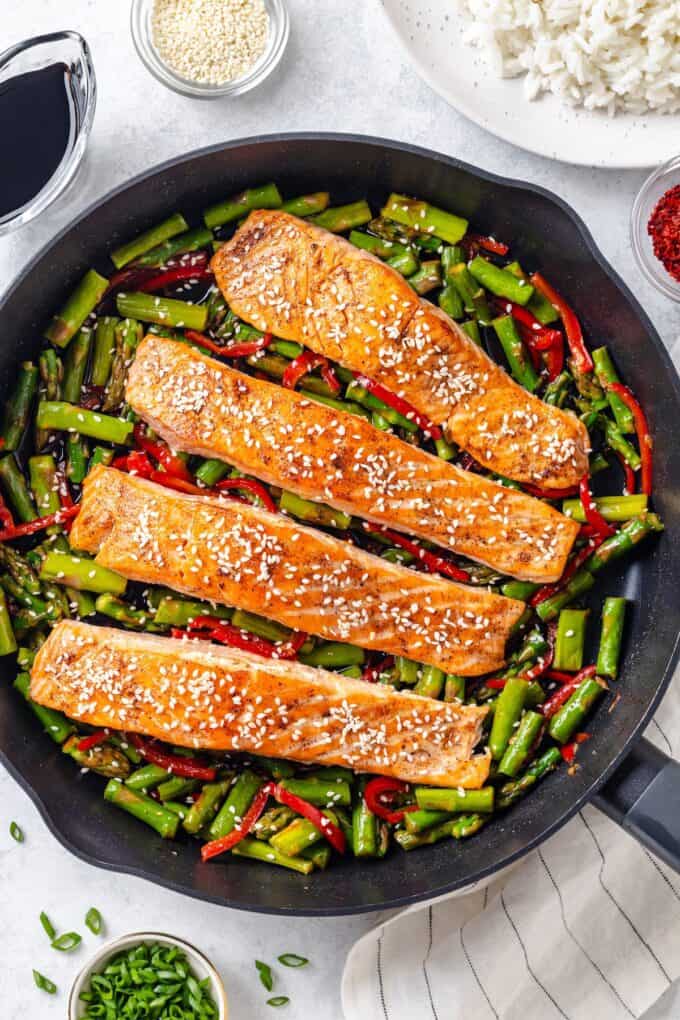 A large dark skillet with seared salmon filets resting on top of a mixture of asparagus and red bell pepper with sesame seeds sprinkled over everything.