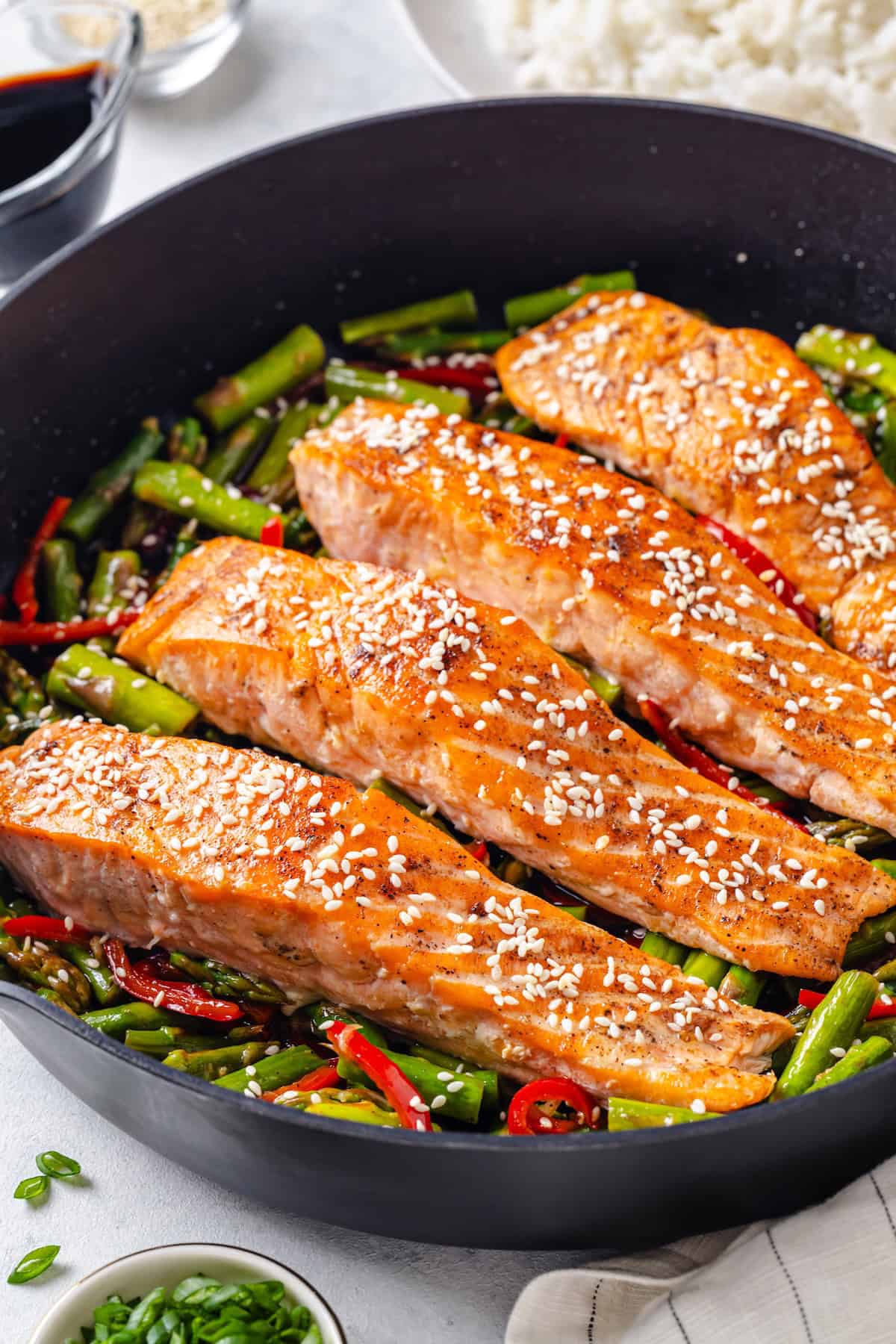 Close up of seared salmon filets in a large dark skillet on a bed of asparagus and red bell pepper pieces.