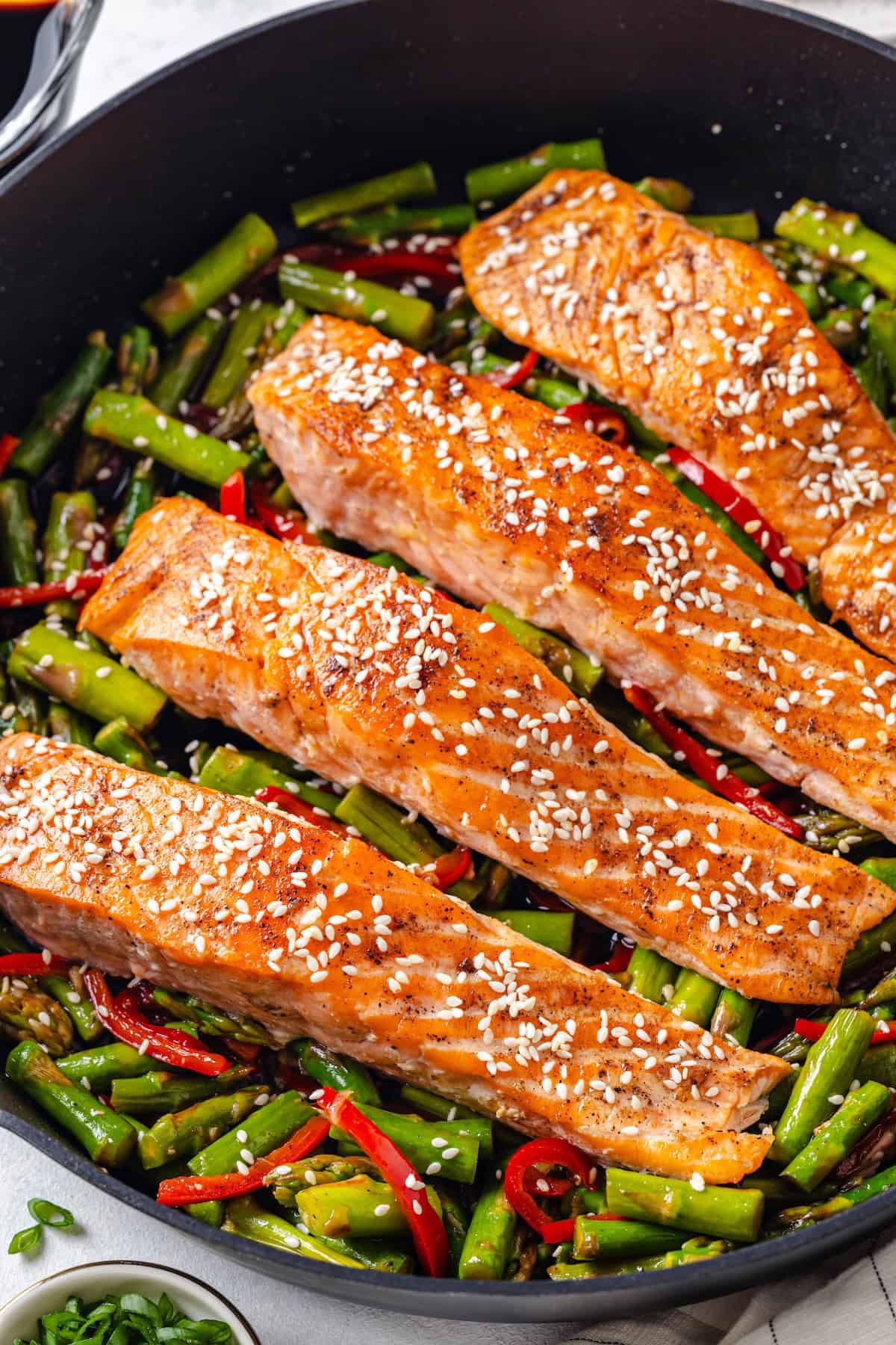 Close up of seared salmon filets topped with sesame seeds in a large dark skillet on a bed of asparagus and red bell pepper pieces.