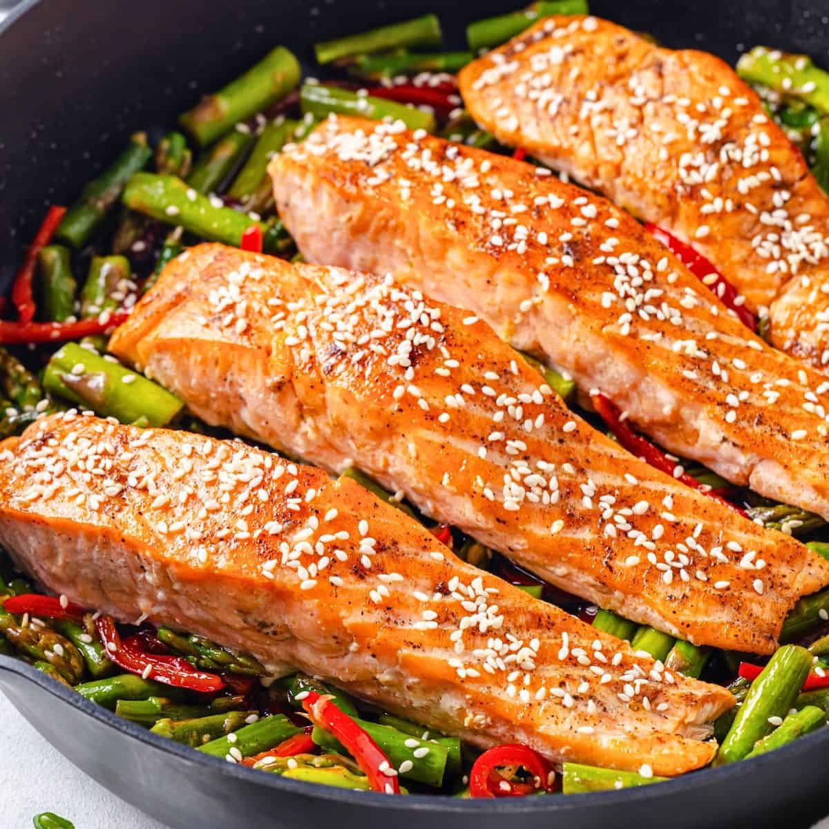 Seared salmon filets in a large dark skillet on a bed of asparagus and red bell pepper pieces with sesame seeds on top.