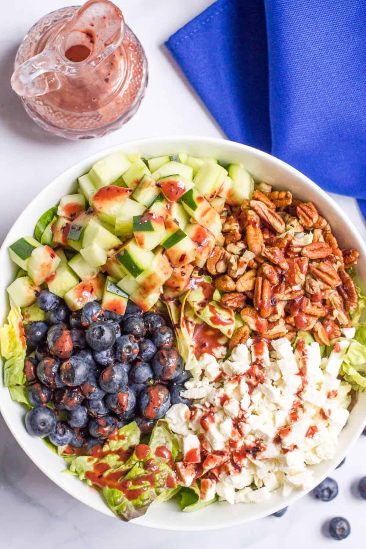 A white serving bowl with a summer salad with blueberries, cucumber, pecans and feta cheese in separate piles with a blueberry vinaigrette drizzled on top and the rest of the glass jar of dressing to the side.