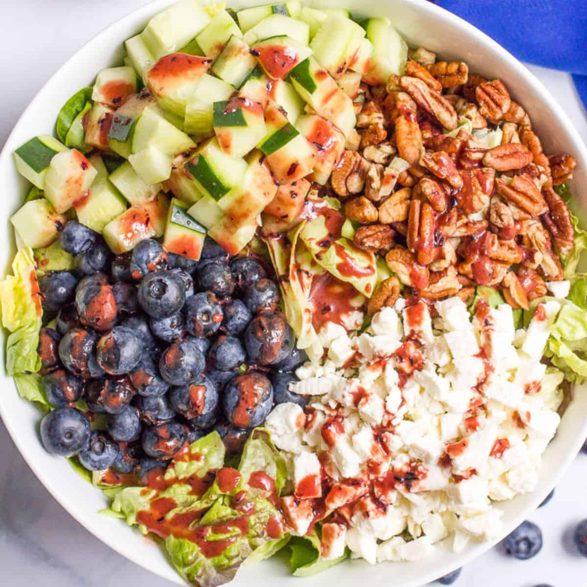A white serving bowl with a summer salad with blueberries, cucumber, pecans and feta cheese in separate piles with a blueberry vinaigrette drizzled on top.
