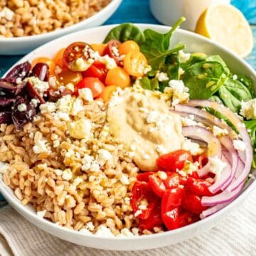 Close up of a low white bowl filled with cooked farro, baby spinach, piles of fresh tomatoes and olives plus red onion and peppers and a dollop of hummus in the middle with feta cheese sprinkled on top.