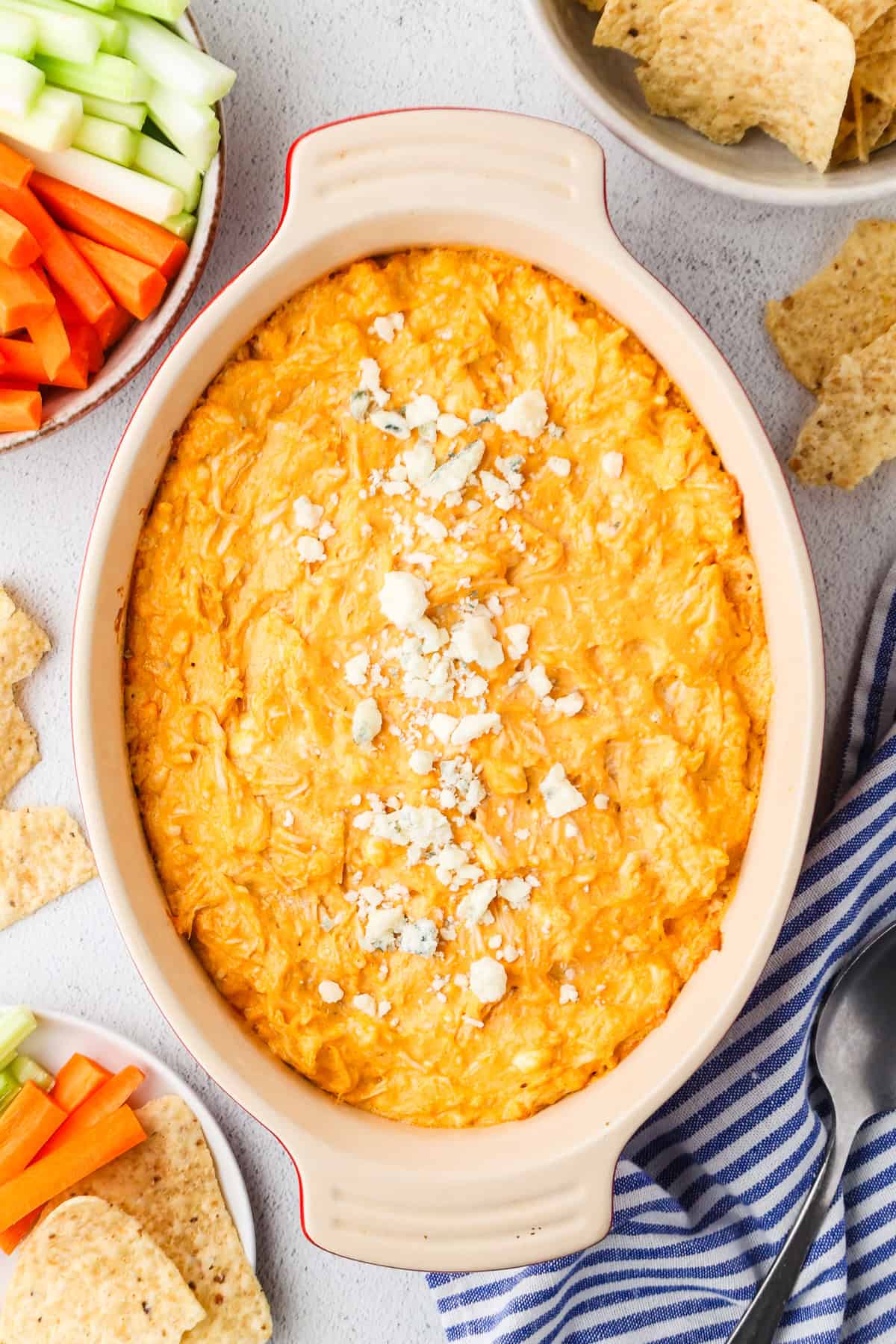 A baked buffalo chicken dip with blue cheese crumbles on top and chips and veggie sticks nearby for dipping.