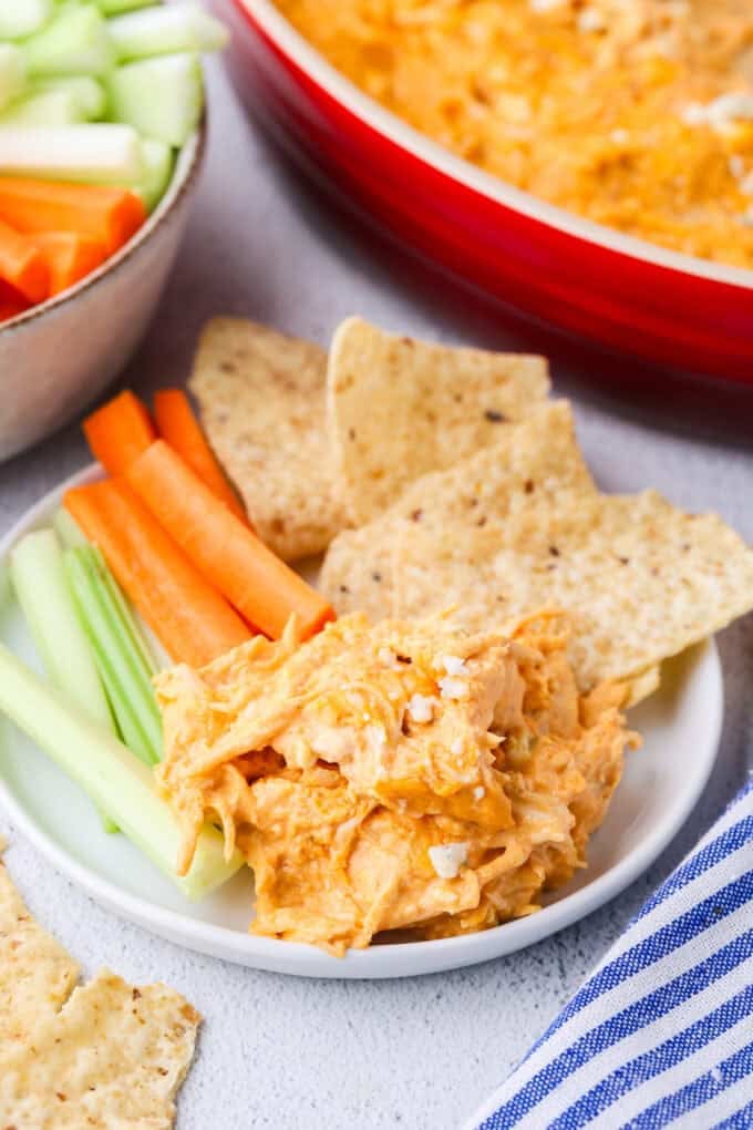 A small white plate with a scoop of a buffalo chicken dip alongside tortilla chips and carrot and celery sticks.