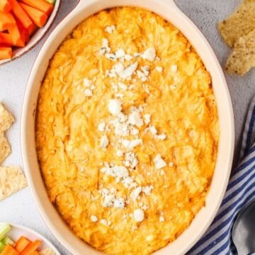 Close up of a baked buffalo chicken dip with blue cheese crumbles on top and chips and veggie sticks nearby for dipping.