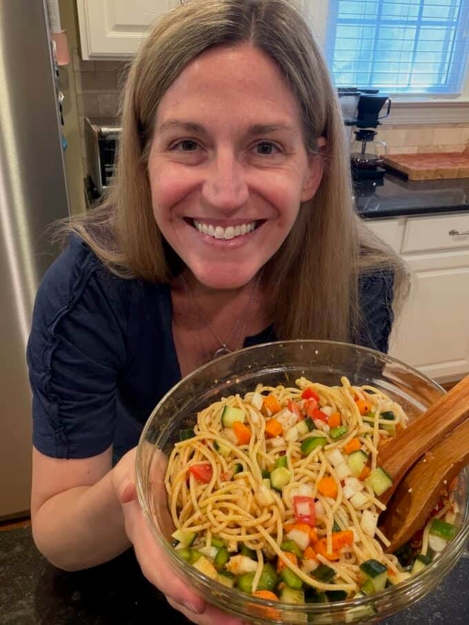 A woman holding a glass bowl full of a cold spaghetti salad with veggies.
