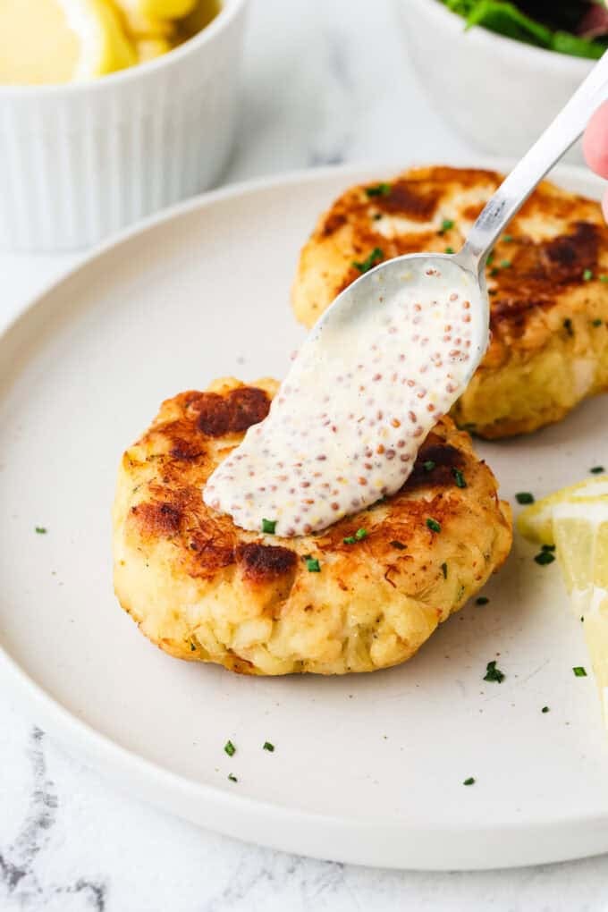 A mustard remoulade sauce being spooned over a crab cake on a white plate.