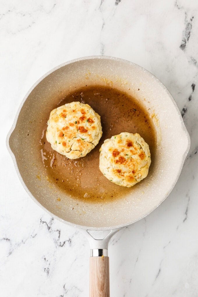 Two crab cakes being cooked in butter in a pan.
