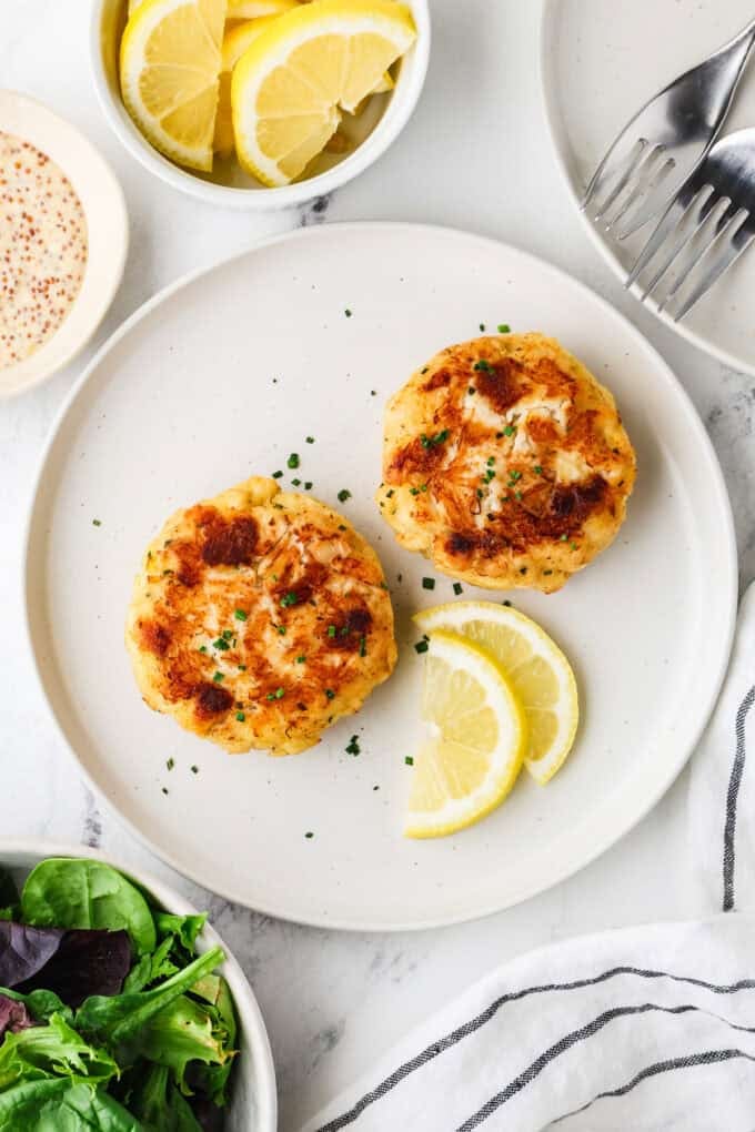 Two cooked, golden brown crab cakes on a white plate with lemon slices to the side and chives sprinkled on top.