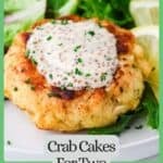Close up of a crab cake with mustard remoulade on top sprinkled with finely chopped chives with a green border and text around the photo.