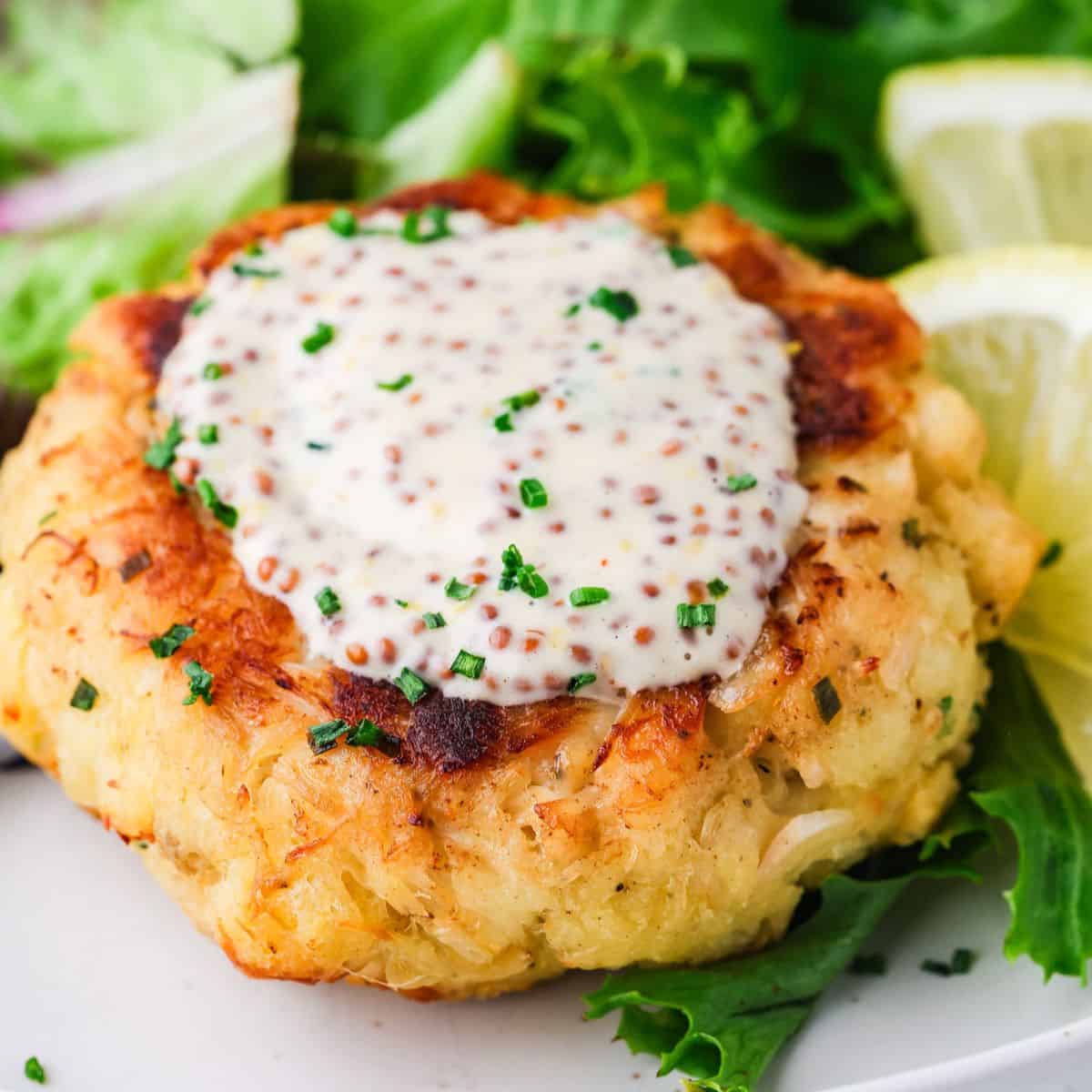 Close up of a crab cake with mustard remoulade on top sprinkled with finely chopped chives with lemon slices to the side.