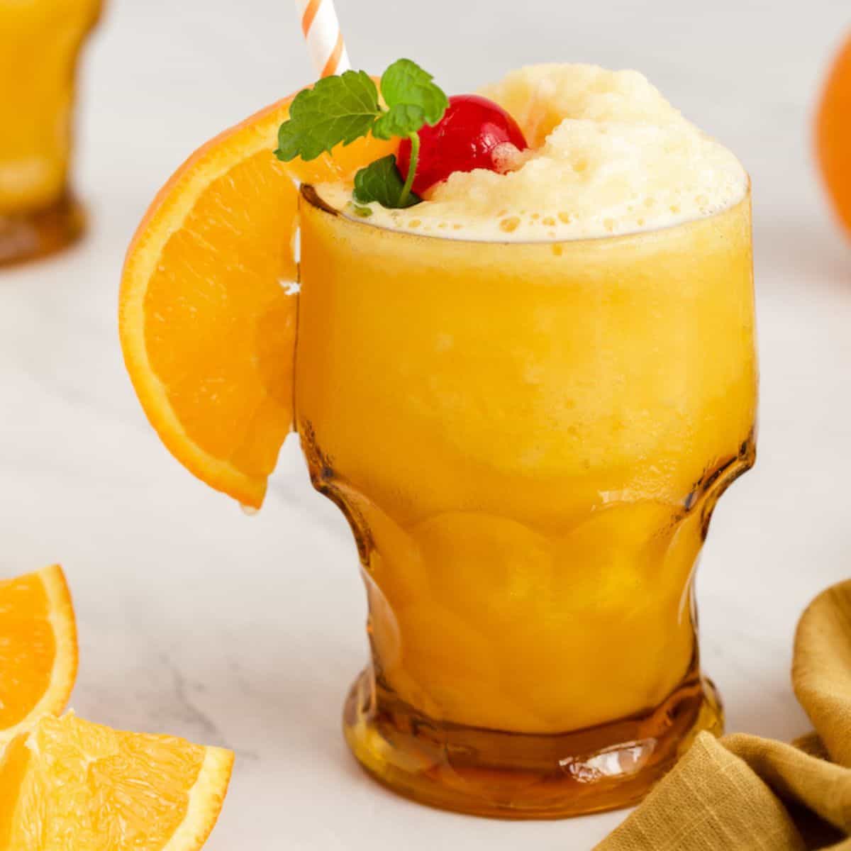 Close up of a slushy frozen screwdriver drink in an amber glass with a cherry and sprig of mint on top plus a straw and an orange slice for garnish.