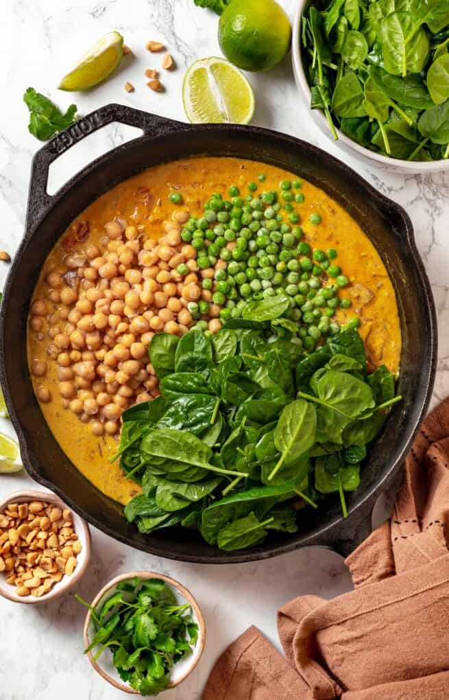 Piles of chickpeas, green peas and fresh spinach being added to a curry sauce in a cast iron pan.