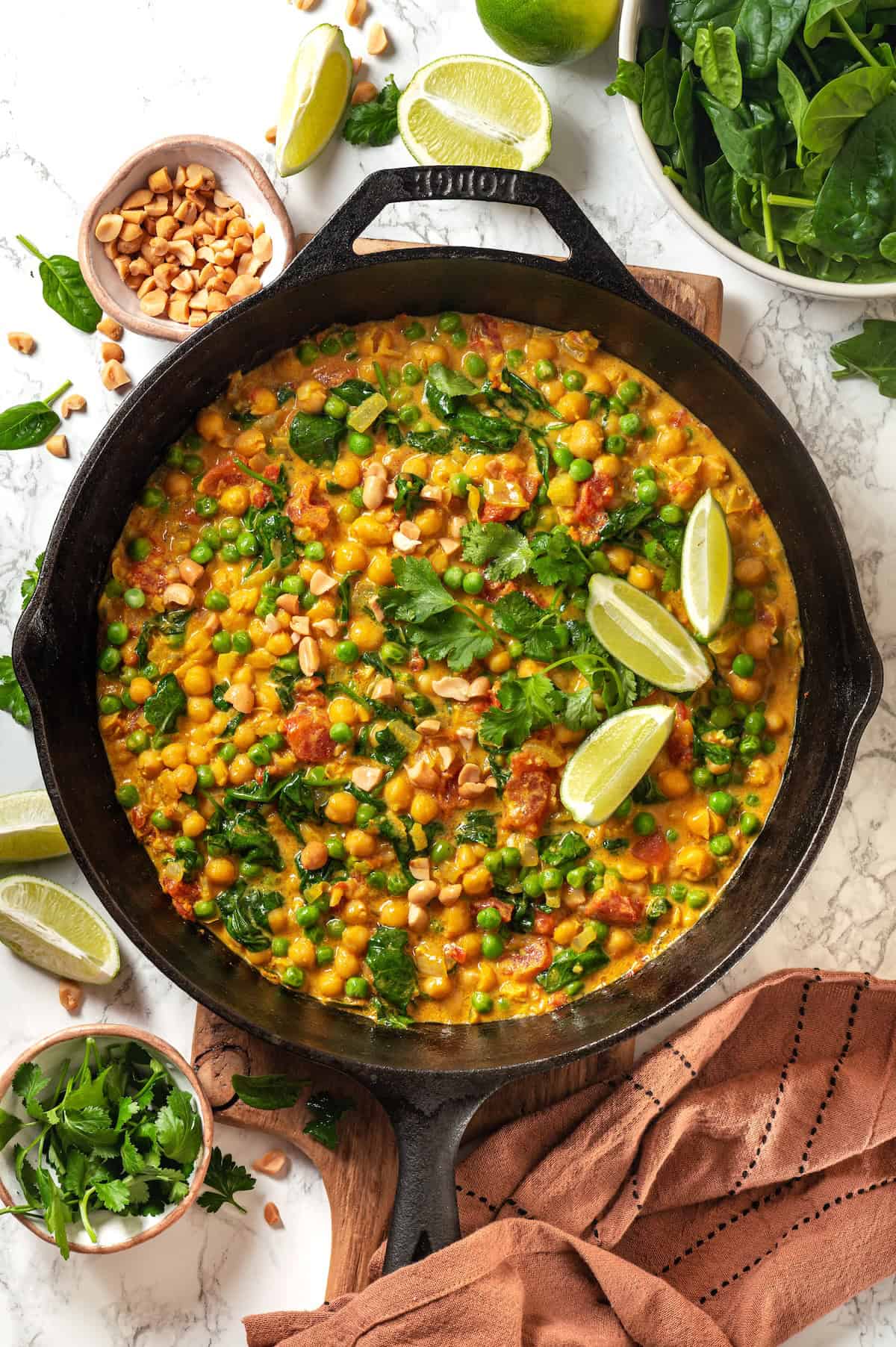 A cast iron pan with a vegetarian curry with chickpeas, spinach, tomatoes and peas with cilantro and lime wedges on top.