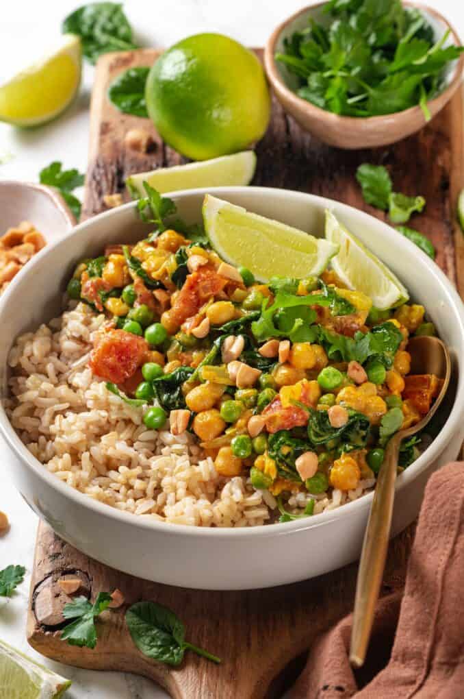 A white bowl with steamed rice topped with a vegetarian chickpea curry with tomatoes and green peas and some lime wedges to the side as garnish.