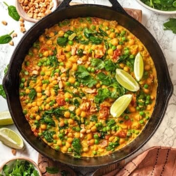 Close up of a cast iron pan with a vegetarian curry with chickpeas, spinach, tomatoes and peas with cilantro and lime wedges on top.