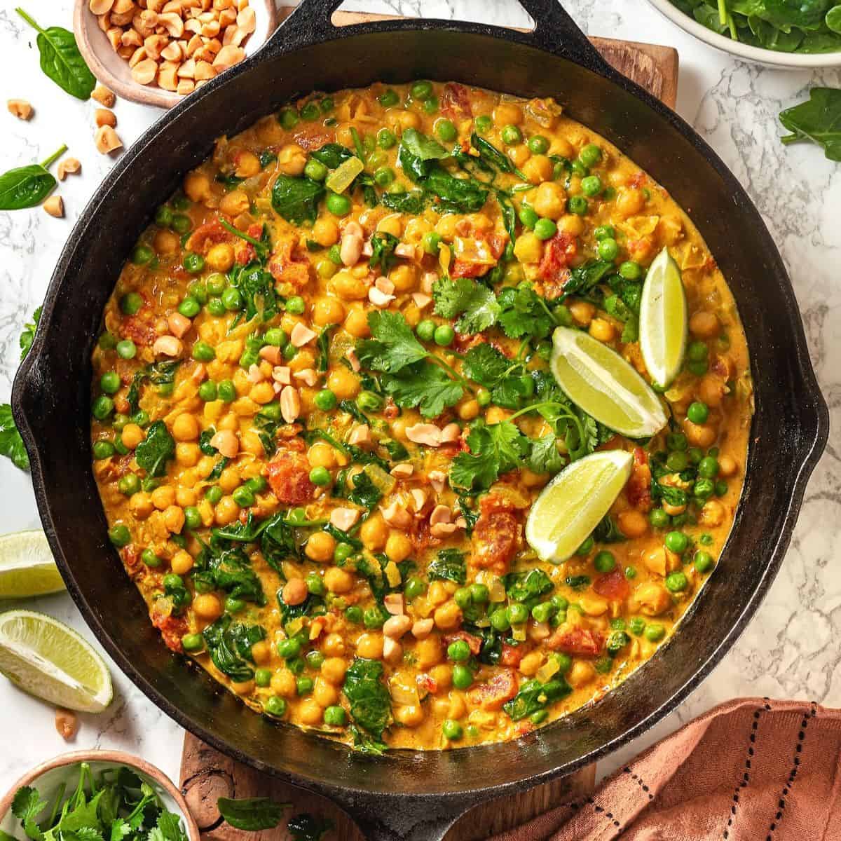Close up of a cast iron pan with a vegetarian curry with chickpeas, spinach, tomatoes and peas with cilantro and lime wedges on top.