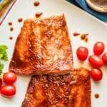Two BBQ slathered filets of salmon on a white platter with splatters of BBQ sauce around and garnishes of fresh herbs and cherry tomatoes.