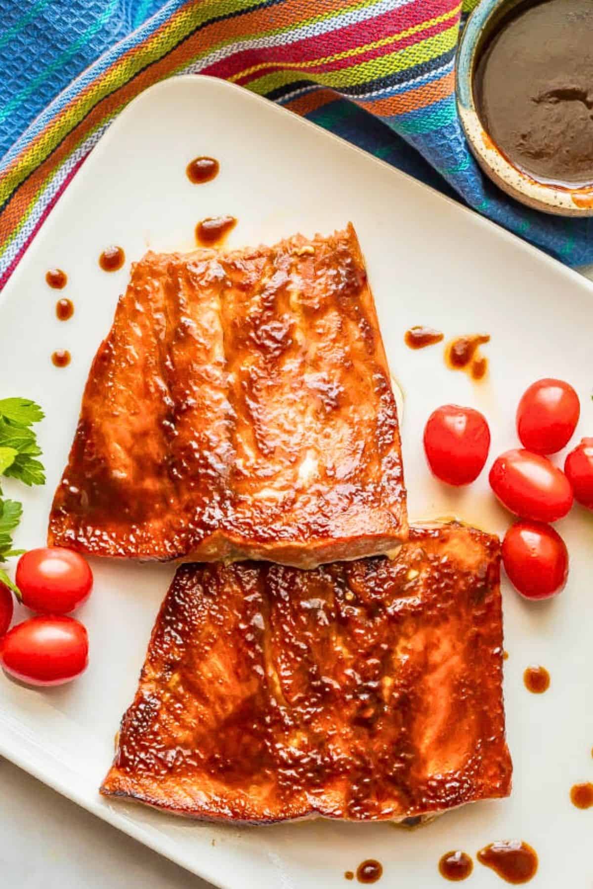 Two BBQ slathered filets of salmon on a white platter with splatters of BBQ sauce around and garnishes of fresh herbs and cherry tomatoes.