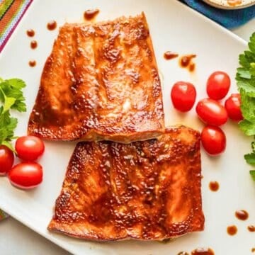 Close up of 2 BBQ slathered filets of salmon on a white platter with splatters of BBQ sauce around and garnishes of fresh herbs and cherry tomatoes.