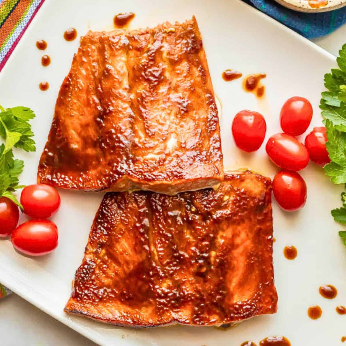 Close up of 2 BBQ slathered filets of salmon on a white platter with splatters of BBQ sauce around and garnishes of fresh herbs and cherry tomatoes.