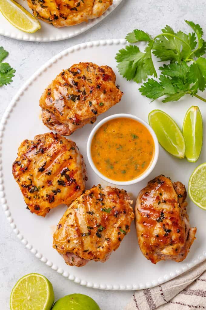Grilled chicken thighs arranged on a white plater with a sauce in the middle and lime wedges scattered about.