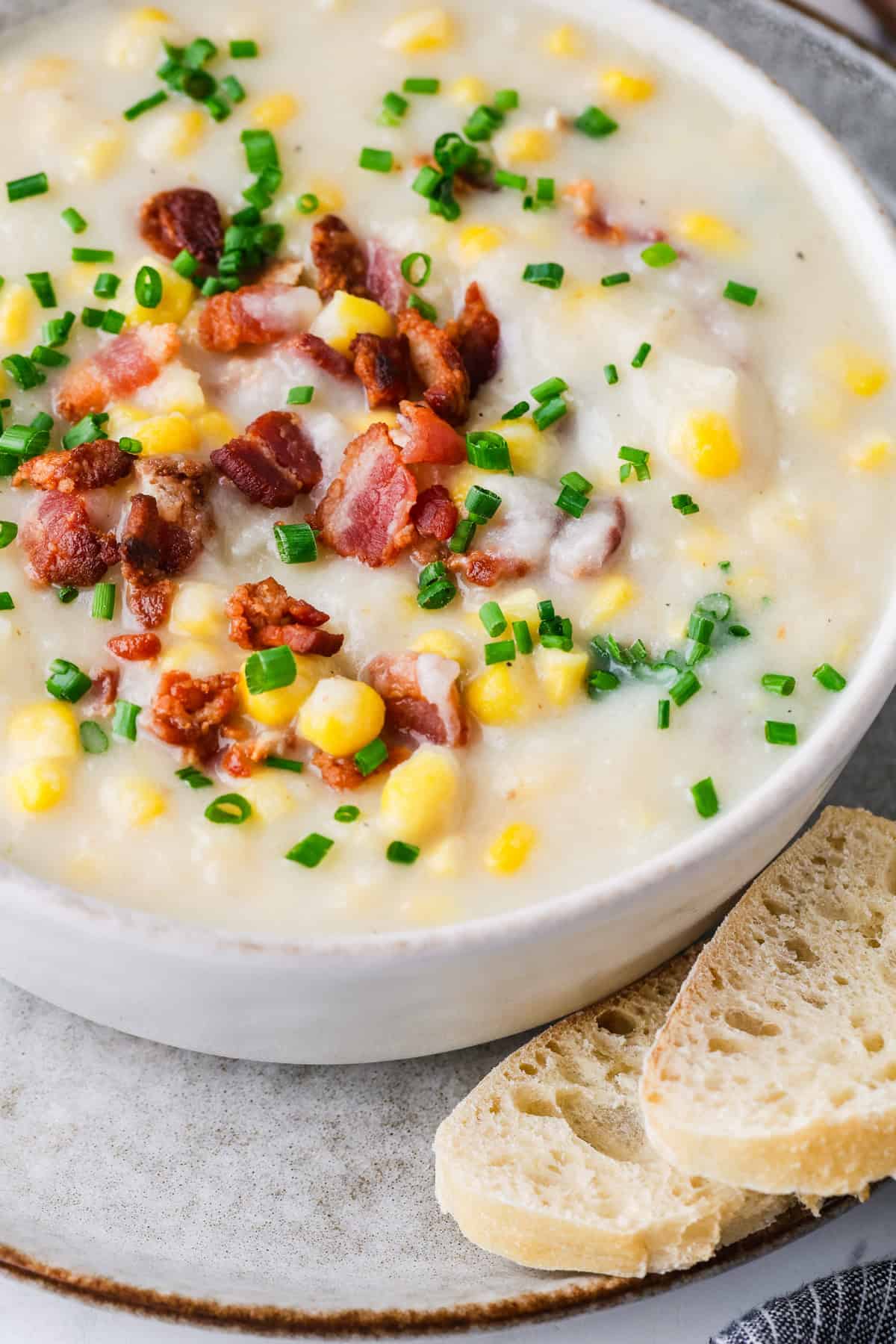 A large white soup bowl full of corn chowder topped with cooked, crisp bacon and chopped chives.