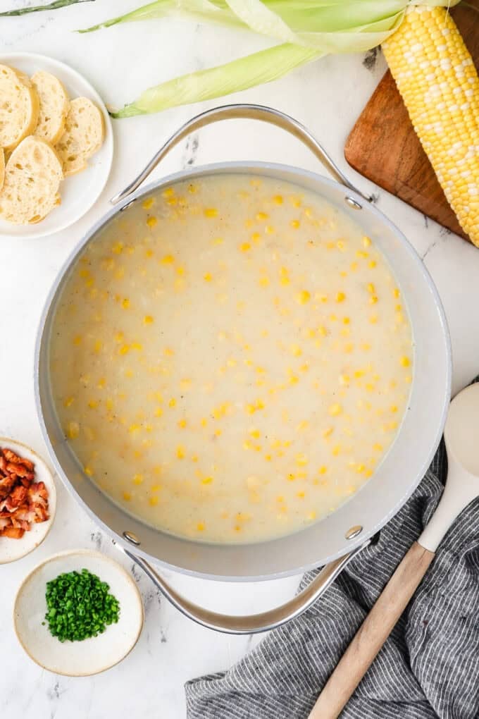 Homemade corn chowder in a large stock pot with toppings to the side and a fresh ear of corn on a cutting board nearby.