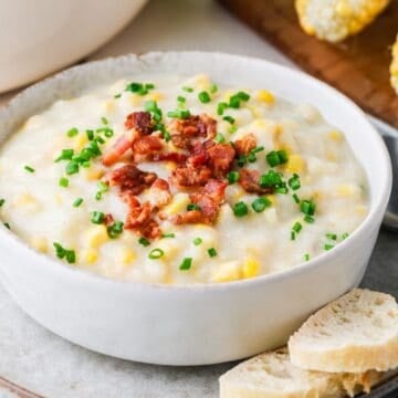 Close up of a bowl full of corn chowder topped with bacon and chives on a plate with bread slices to the side.
