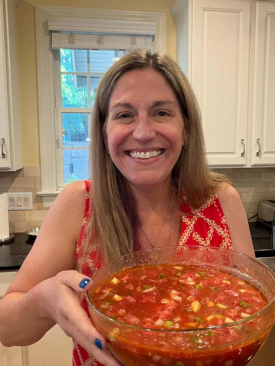 A woman holding a large glass bowl full of homemade gazpacho soup with fresh veggies.