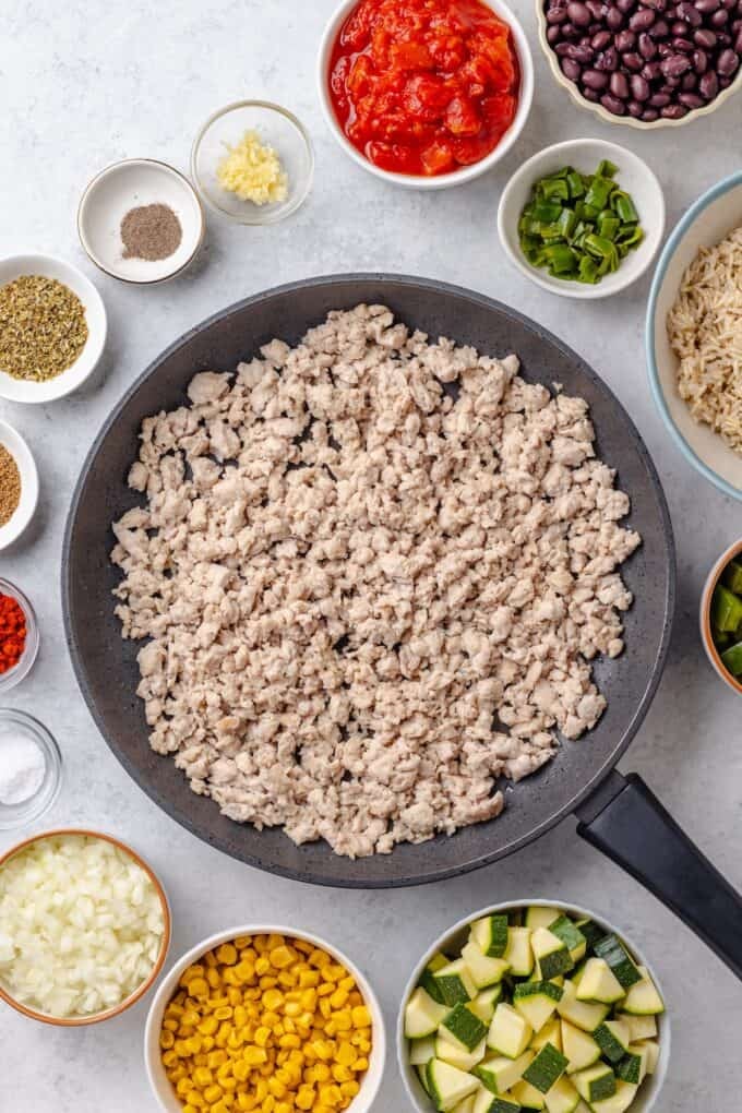 Cooked, crumbled ground turkey in a large skillet with bowls of other ingredients around it to be added in.