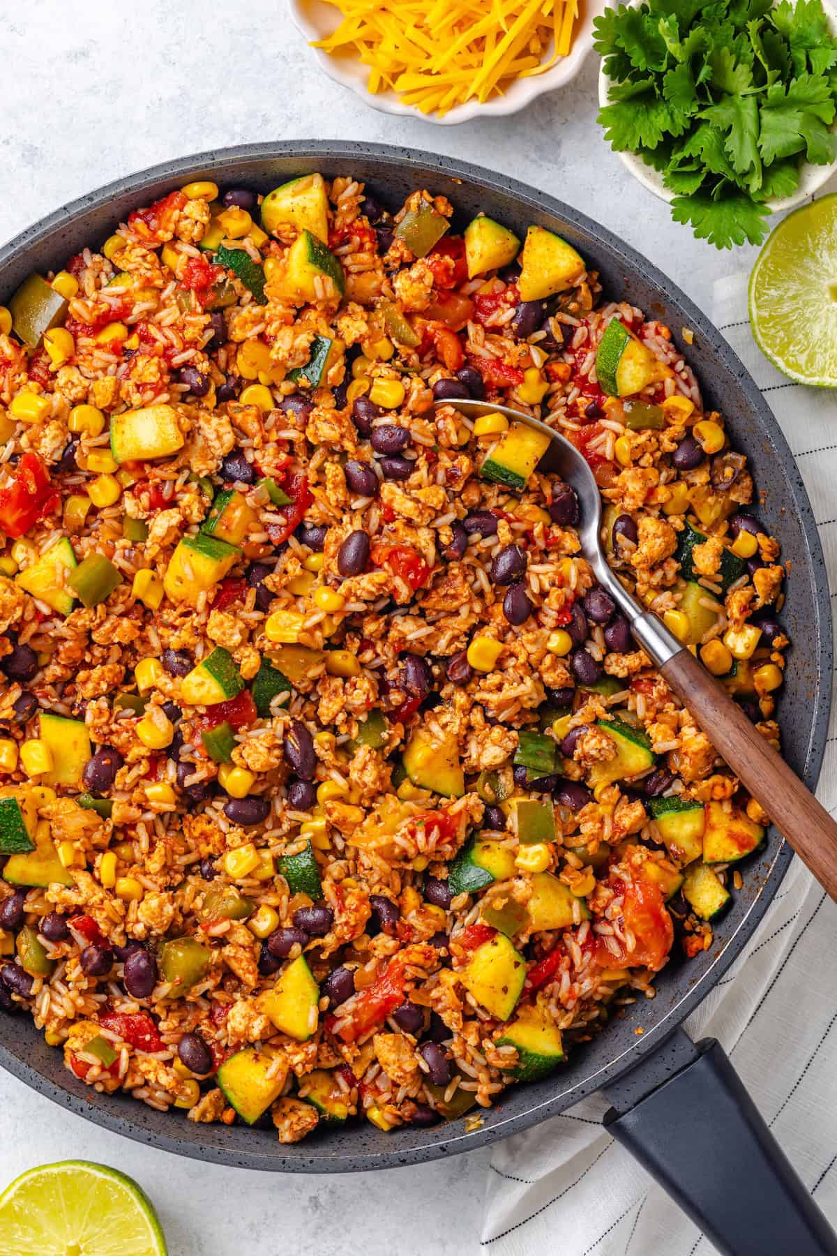 A spoon resting in a colorful skillet of ground meat with rice, beans and veggies.