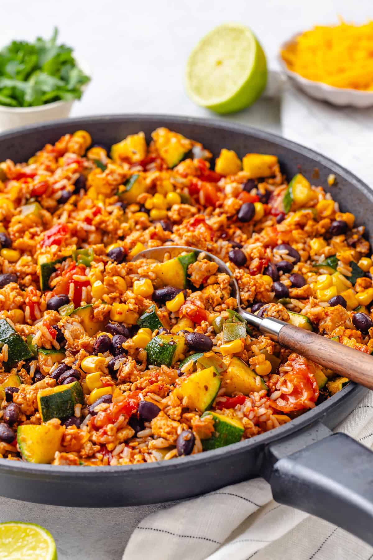 A spoon resting in a pan full of a rice, ground meat, black bean, zucchini and tomato skillet.