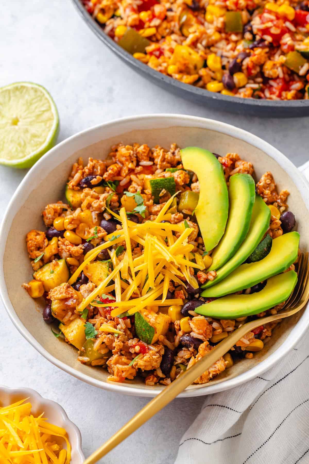 A bowl full of a ground turkey and rice mixture with beans and veggies, topped with avocado slices and shredded cheddar cheese.